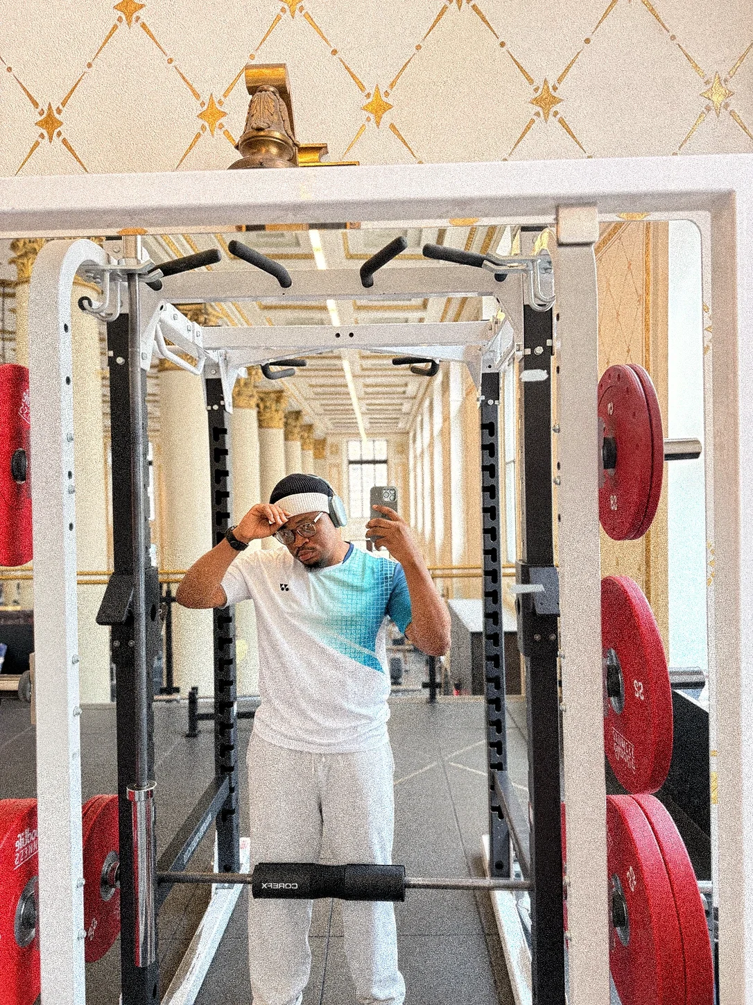 Oluwayemi taking a mirror selfie in a gym, standing in a squat rack with red weight plates